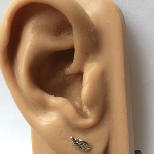 May include: A silver ear cuff with a textured design in the shape of a wing. The cuff is shown on a model ear.