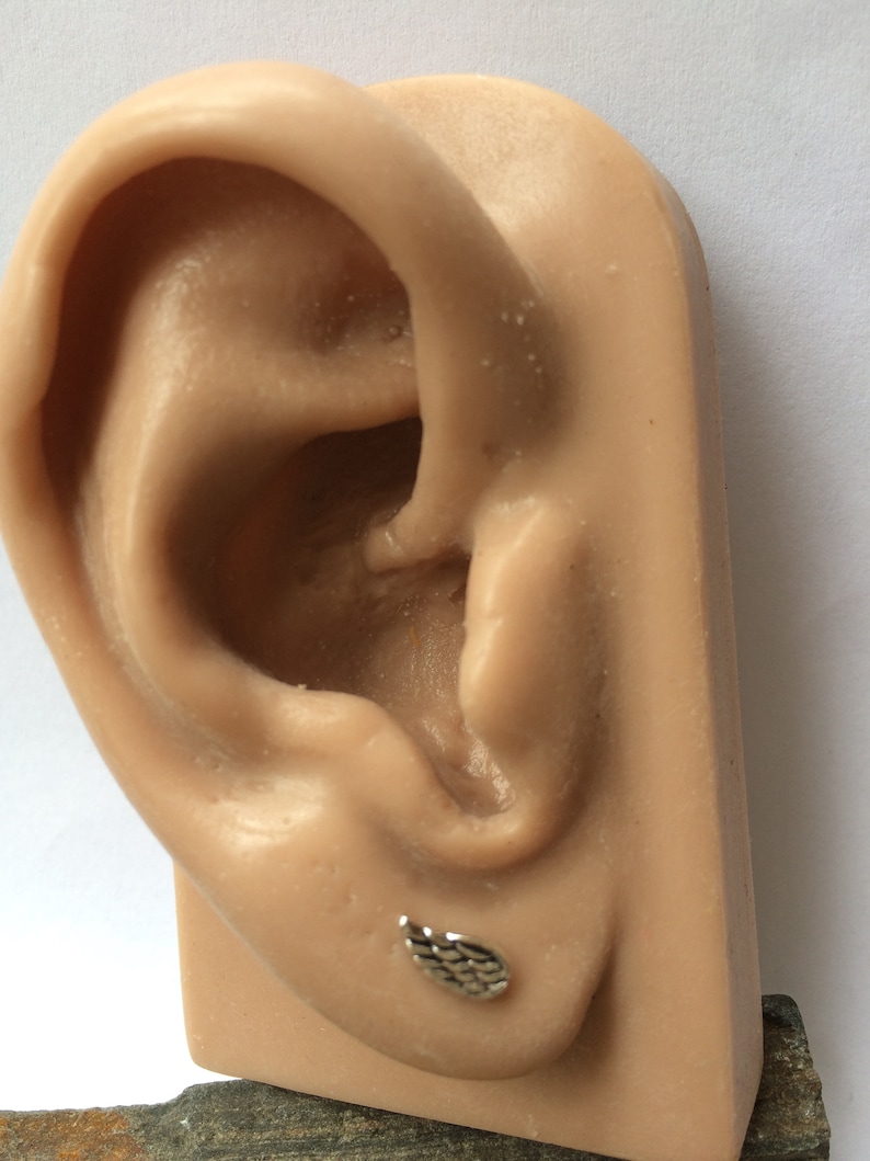 May include: A silver ear cuff with a textured design in the shape of a wing. The cuff is shown on a model ear.