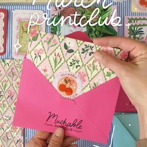 Könnte beinhalten: Ein rosa Umschlag mit einem floral gemusterten Einsatz und einem Kirschaufkleber wird geöffnet, um den Inhalt zu zeigen. Die Worte "March printclub" sind in weißer Schrift geschrieben. Der Umschlag trägt die Aufschrift "Muchable Print Club".