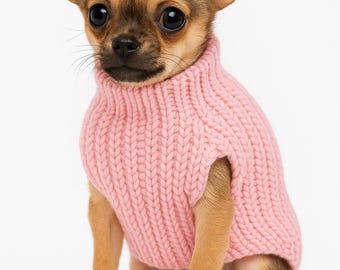 Puede incluir: Un cachorro de chihuahua pequeño, marrón y canela, con un suéter de cuello alto de punto grueso rosa claro. El perro está sentado y mirando directamente a la cámara sobre un fondo blanco. El suéter cubre el cuerpo del perro.