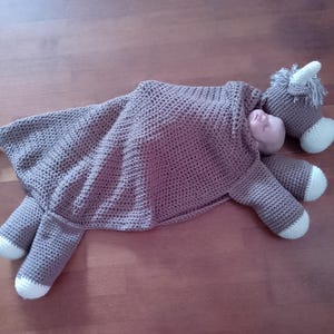 May include: A brown crocheted horse blanket with a white trim. The blanket is shaped like a horse and has a hood. The blanket is lying on a wooden floor.