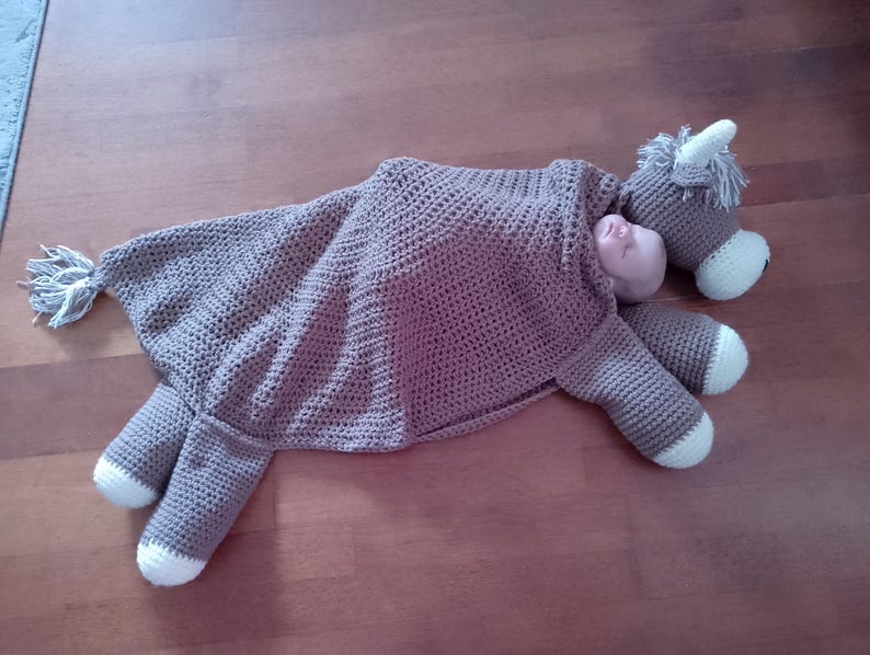 May include: A brown crocheted horse blanket with a white trim. The blanket is shaped like a horse and has a hood. The blanket is lying on a wooden floor.