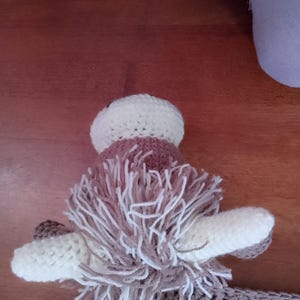 May include: A brown and white crocheted stuffed animal with long, shaggy hair. The animal has large, white ears and a brown nose.