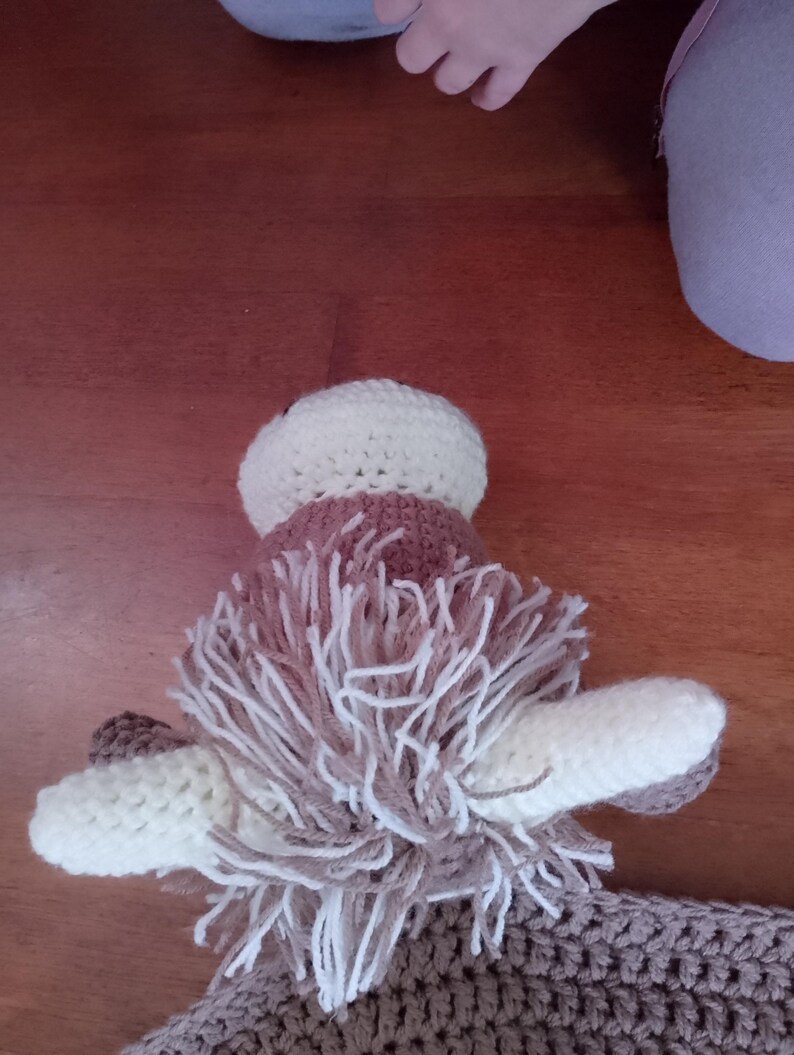 May include: A brown and white crocheted stuffed animal with long, shaggy hair. The animal has large, white ears and a brown nose.