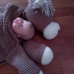 May include: A brown crocheted unicorn stuffed animal with white accents. The unicorn is lying on its side with its head resting on a baby doll.