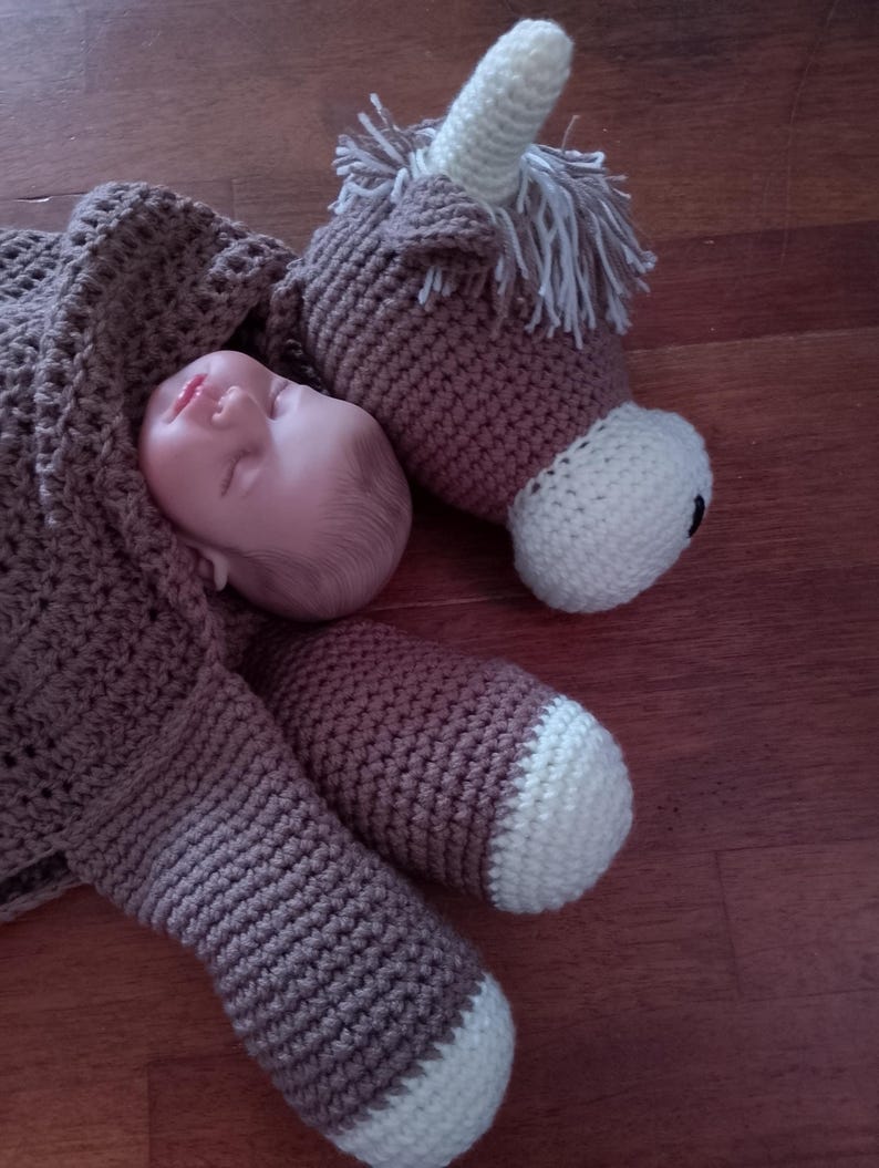 May include: A brown crocheted unicorn stuffed animal with white accents. The unicorn is lying on its side with its head resting on a baby doll.