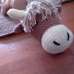 May include: A brown and white crocheted stuffed animal resembling a Highland cow with a long, shaggy mane. The cow is lying on a brown surface.