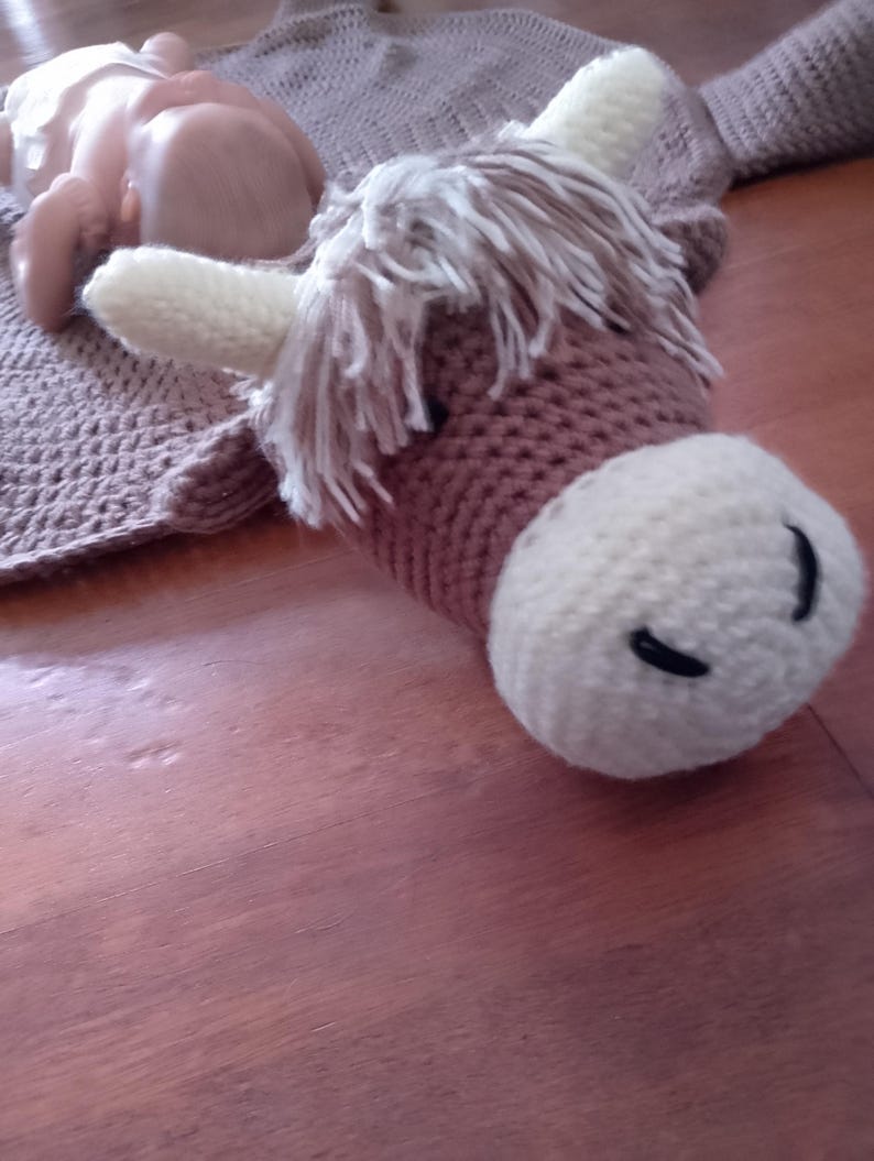 May include: A brown and white crocheted stuffed animal resembling a Highland cow with a long, shaggy mane. The cow is lying on a brown surface.