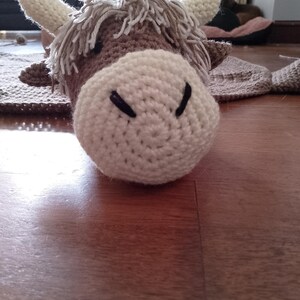 May include: A crocheted brown and white cow with a fluffy mane. The cow is lying on a wooden floor.
