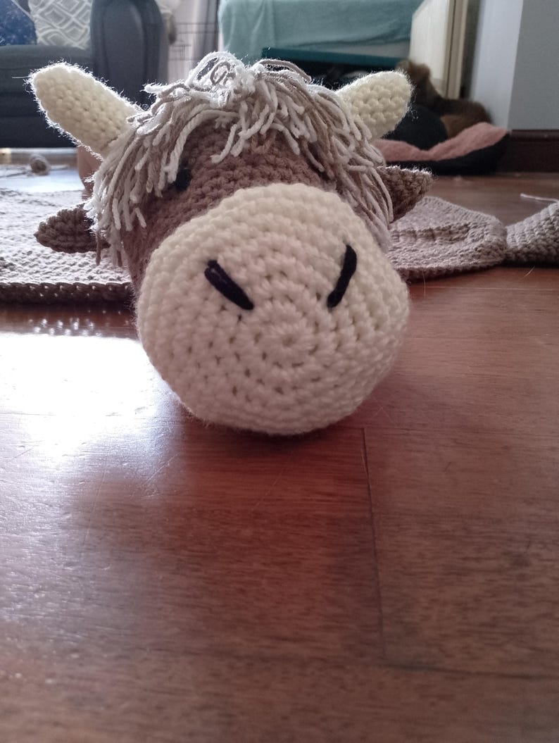 May include: A crocheted brown and white cow with a fluffy mane. The cow is lying on a wooden floor.