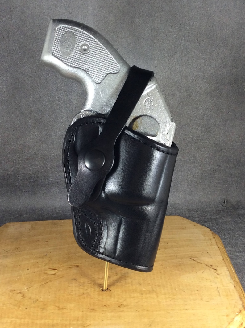 Op de afbeelding: Een zwarte leren holster met een zilveren handvuurwapen. De holster is ontworpen om het vuurwapen veilig vast te houden. De afbeelding toont het pistool gedeeltelijk in de holster gestoken, met een riem die de bovenkant vastzet.