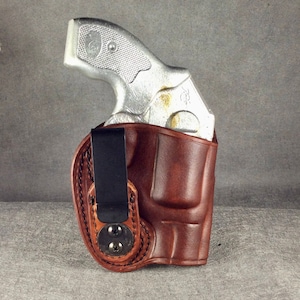 May include: A brown leather holster with a black clip, holding a silver revolver. The holster is designed to fit the gun's shape, with visible stitching and a secure fit. The background is a solid gray.