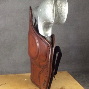 May include: A brown leather holster with a silver gun inside. The holster has a belt loop and is designed to be worn on the hip.