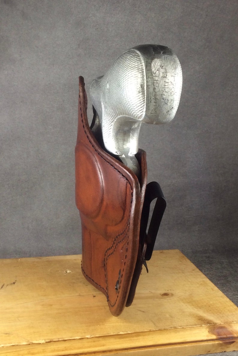 May include: A brown leather holster with a silver gun inside. The holster has a belt loop and is designed to be worn on the hip.