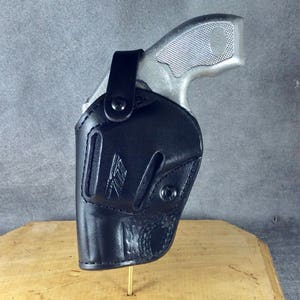 Op de afbeelding: Een zwart leren holster met een zilverkleurig handvuurwapen. De holster heeft een gestikt ontwerp en een druksluiting. De greep en trekker van het handvuurwapen zijn zichtbaar. De holster wordt gepresenteerd op een houten oppervlak.