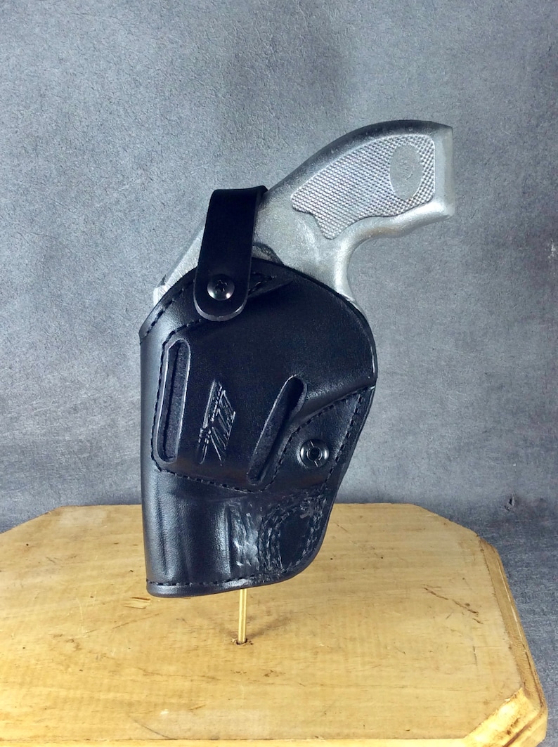 Op de afbeelding: Een zwart leren holster met een zilverkleurig handvuurwapen. De holster heeft een gestikt ontwerp en een druksluiting. De greep en trekker van het handvuurwapen zijn zichtbaar. De holster wordt gepresenteerd op een houten oppervlak.