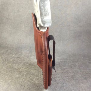 May include: A brown leather holster with a silver metal object inside. The holster has a black belt clip.