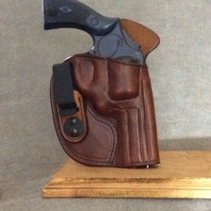 May include: A brown leather holster with a black clip and a silver screw. The holster is designed to hold a small revolver. The revolver is blue and has a wooden grip.