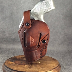 Op de afbeelding: Een bruin leren holster met een zilveren handvuurwapen. De holster is gemaakt van gestikt leer met zwarte klinknagels en een druksluiting. Het handvuurwapen is gedeeltelijk zichtbaar, met een getextureerde grip. De holster wordt op een houten standaard getoond.