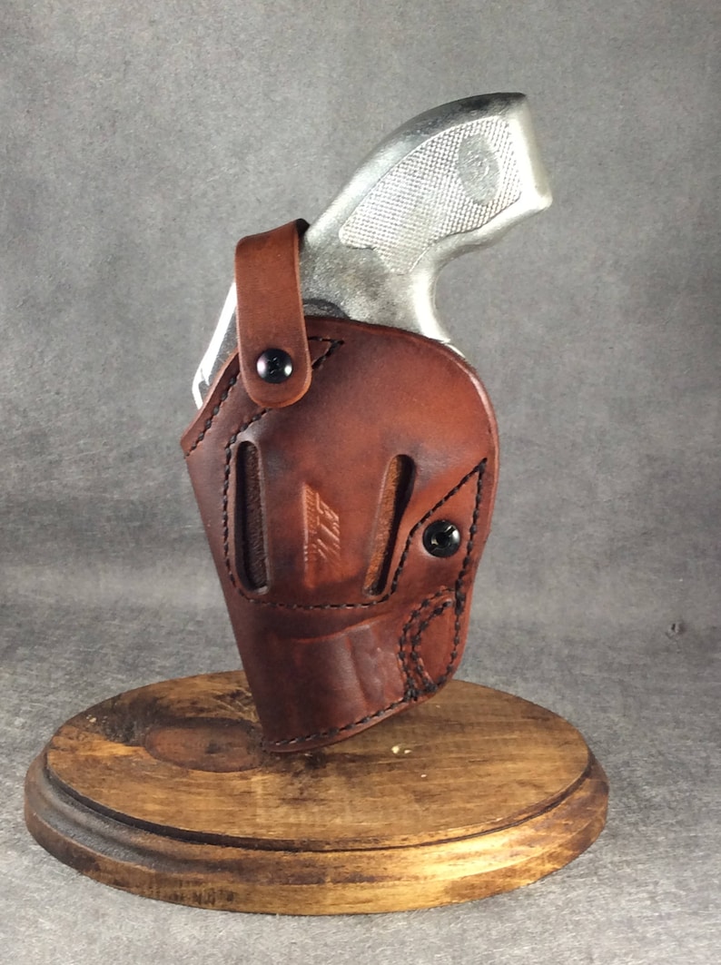 Op de afbeelding: Een bruin leren holster met een zilveren handvuurwapen. De holster is gemaakt van gestikt leer met zwarte klinknagels en een druksluiting. Het handvuurwapen is gedeeltelijk zichtbaar, met een getextureerde grip. De holster wordt op een houten standaard getoond.