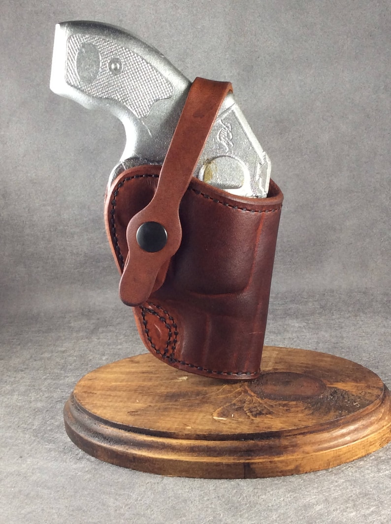 Op de afbeelding: Een bruin leren holster met een zilveren pistool erin. Het holster heeft een riem met een zwarte knoopsluiting. Het holster staat op een houten standaard met een cirkelvormige basis. De achtergrond is effen grijs.