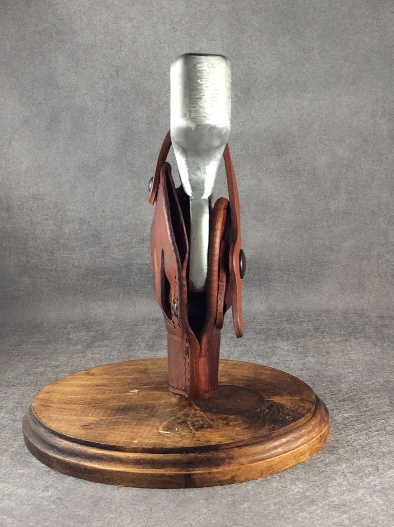 Op de afbeelding: Een handgemaakte flesopener met een bruin leren foedraal en een houten voet. De metalen opener is zilverkleurig en de leren foedraal is gestikt. De houten voet is rond en donkerbruin gebeitst.