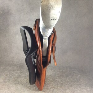 May include: A brown leather holster with a silver gun inside. The holster has a black belt clip attached to it.