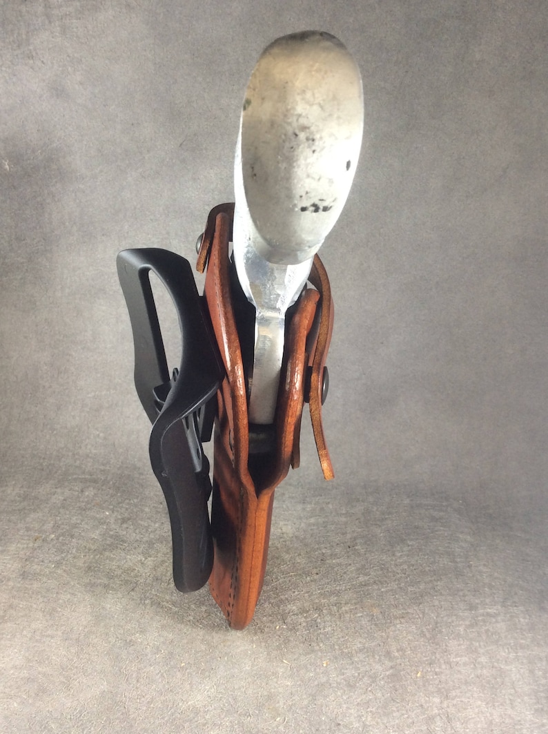 May include: A brown leather holster with a silver gun inside. The holster has a black belt clip attached to it.