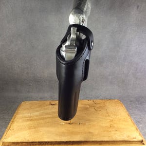 May include: A black leather holster for a silver revolver. The holster is designed to be worn on the hip and has a belt loop. The revolver is partially visible in the holster.
