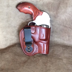 Könnte beinhalten: Ein Revolver aus Edelstahl mit Holzmaserung ist teilweise in einem braunen Lederholster untergebracht. Das Holster hat einen schwarzen Metallclip und ist mit schwarzem Faden genäht. Der Revolver ist eine kleine, kompakte Schusswaffe.