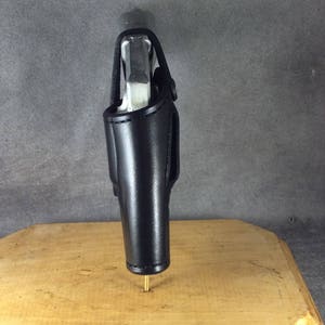 Op de afbeelding: Zwarte leren holster met een zilveren pistool erin. De holster is ontworpen om een vuurwapen te bevatten en heeft een riemlus. De holster wordt gepresenteerd op een houten oppervlak.