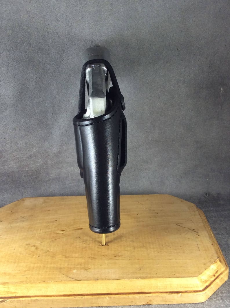 Op de afbeelding: Zwarte leren holster met een zilveren pistool erin. De holster is ontworpen om een vuurwapen te bevatten en heeft een riemlus. De holster wordt gepresenteerd op een houten oppervlak.