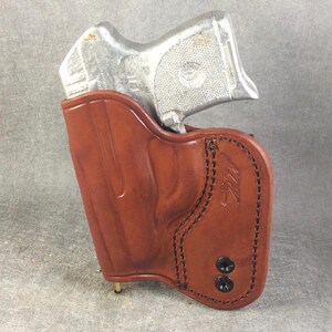 May include: A brown leather holster with a silver handgun inside. The holster has a stitched design and two black snaps.