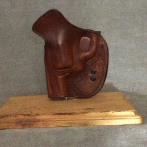 May include: A brown leather holster for a handgun, displayed on a wooden base.