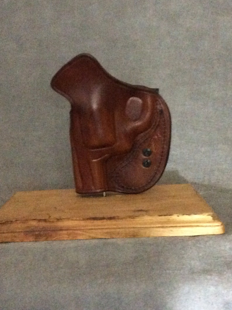 May include: A brown leather holster for a handgun, displayed on a wooden base.