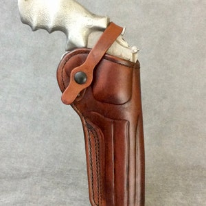 Op de afbeelding: Een bruine leren holster met een gestikt ontwerp en een band. De holster is ontworpen om een ​​zilveren revolver te houden.