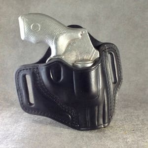 Könnte beinhalten: Ein schwarzes Lederholster für eine kleine silberne Pistole. Das Holster hat eine Gürtelschlaufe und einen Druckknopfverschluss.