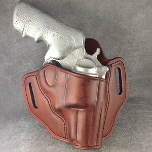 May include: A brown leather holster for a silver revolver. The holster has a belt loop and a strap to secure the gun.