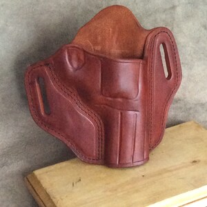 May include: A brown leather holster with stitching detail. The holster has two belt loops and a flap that covers the opening.