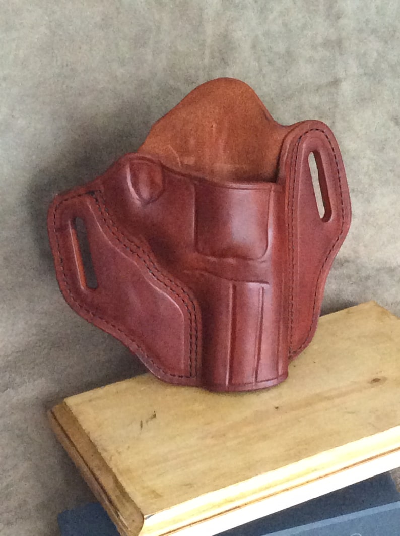 May include: A brown leather holster with stitching detail. The holster has two belt loops and a flap that covers the opening.