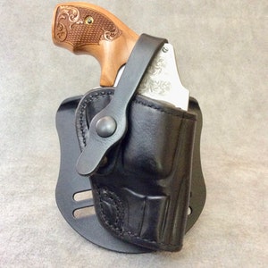 Könnte beinhalten: Ein schwarzes Lederholster mit einem silbernen Revolver darin. Der Revolver hat einen hölzernen Griff mit aufwendigen Schnitzereien. Das Holster hat eine Gürtelschlaufe und einen Druckknopfverschluss.