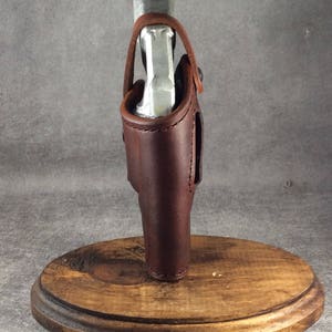 Op de afbeelding: Een bruin leren holster met stiksels, met een zilveren voorwerp. Het holster is gemonteerd op een ronde houten basis. Het leer heeft een rijke, donkere kleur en het ontwerp suggereert een handgemaakt item.
