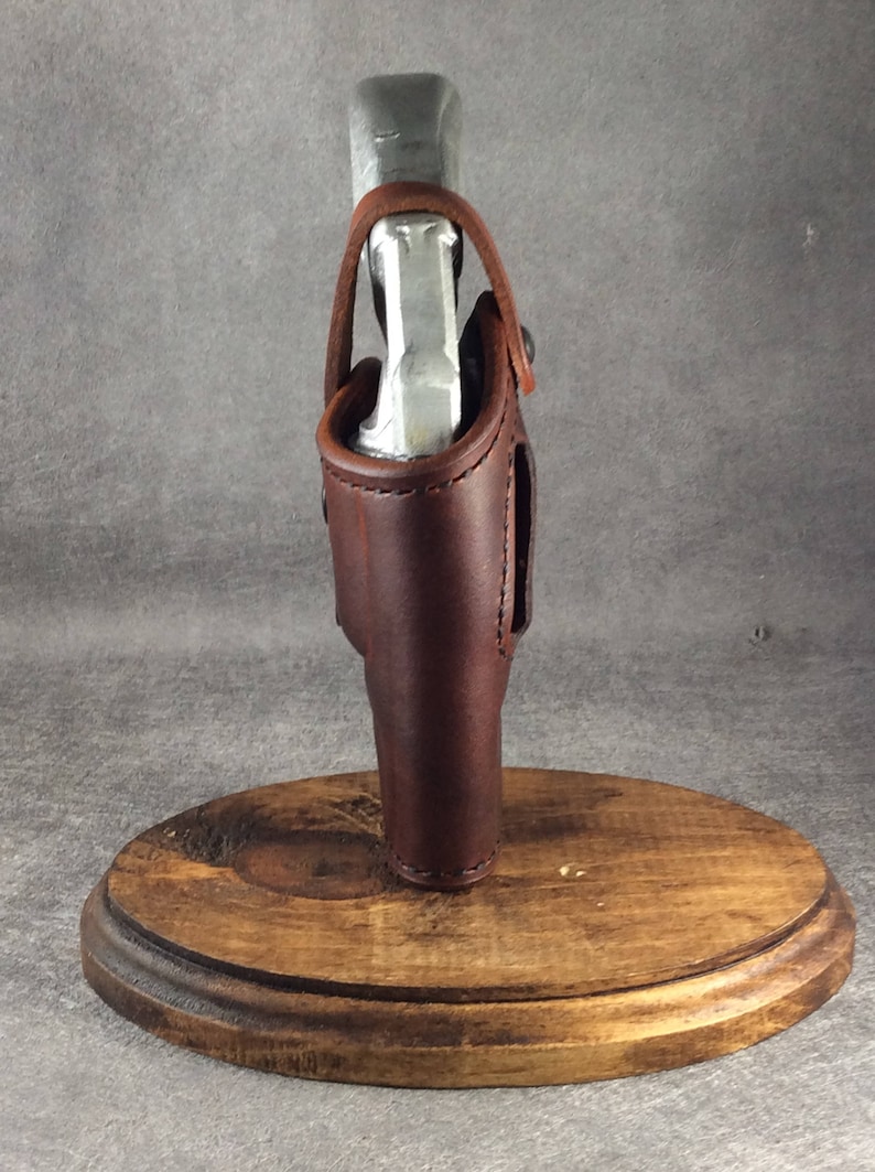 Op de afbeelding: Een bruin leren holster met stiksels, met een zilveren voorwerp. Het holster is gemonteerd op een ronde houten basis. Het leer heeft een rijke, donkere kleur en het ontwerp suggereert een handgemaakt item.