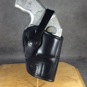 Può includere: Un fodero in pelle nera con un revolver argentato all'interno. Il fodero ha una cinghia e un bottone a pressione.