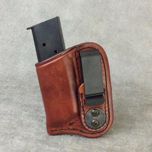 Może przedstawiać: Brązowa skórzana kabura z czarnym klips i magazynkiem w środku. Kabura ma pikowany wzór i dwa śruby z boku.