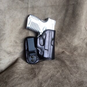 Op de afbeelding: Een zwart leren holster met een zilveren pistool erin. Het holster heeft een clip aan de zijkant om aan een riem te bevestigen.
