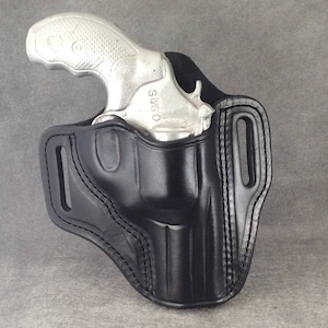Könnte beinhalten: Ein schwarzes Lederholster mit einem silbernen Revolver darin. Das Holster hat eine Gürtelschlaufe und einen Druckknopfverschluss. Der Revolver hat eine silberne Oberfläche und einen schwarzen Griff.