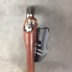May include: A brown leather holster with a silver gun inside. The holster is attached to a black metal belt clip.