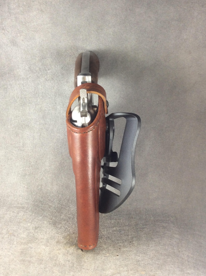 May include: A brown leather holster with a silver gun inside. The holster is attached to a black metal belt clip.