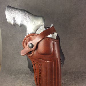 May include: A brown leather holster with a silver handgun inside. The holster has a strap with a snap closure and is designed for concealed carry.